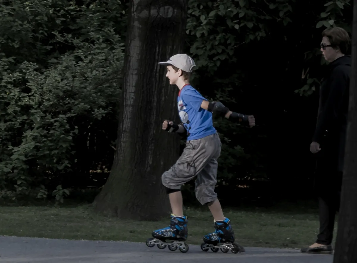 Boy rollerblading through a park while a dark-clad woman watches from behind